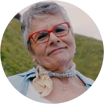 A circular headshot of Marilyn. She is about 55-65 years old. She has short grey hair, red glasses, a blue and yellow scarf tied around her neck and a silver necklace. She is wearing a blue button-up shirt with a white top. In the background is a grassy field.